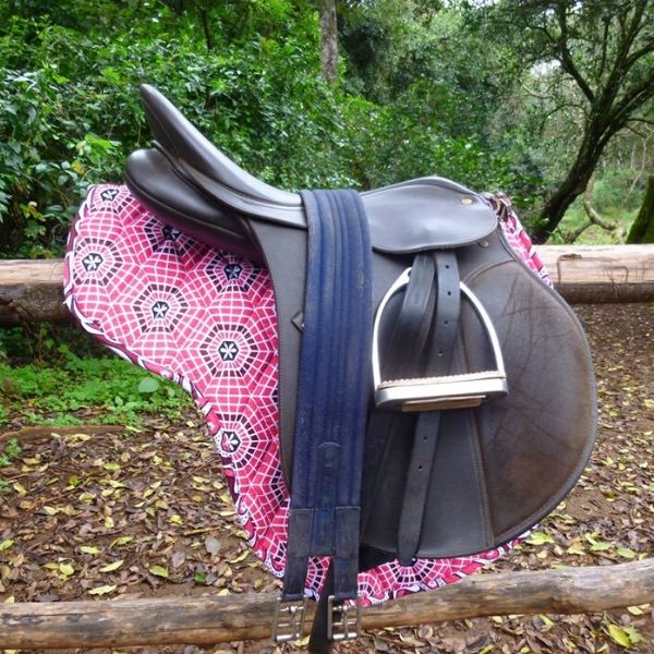 A saddle on a pink kanga numnah resting on a wooden rail in a leafy yard