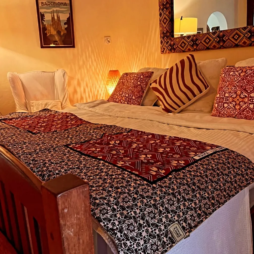 A bed dressed with a red and black kanga throw and patterned cushions in warm lamplight
