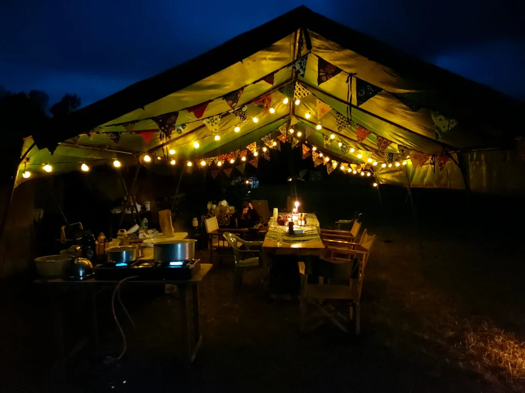 A camp mess tent at night lit by festoon lights and hung with kanga bunting over a candlelit table