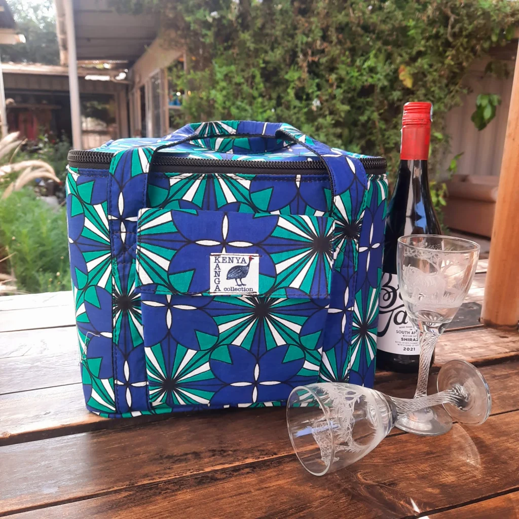 A blue and green kanga cooler bag beside a bottle of red wine and two engraved glasses on a wooden table