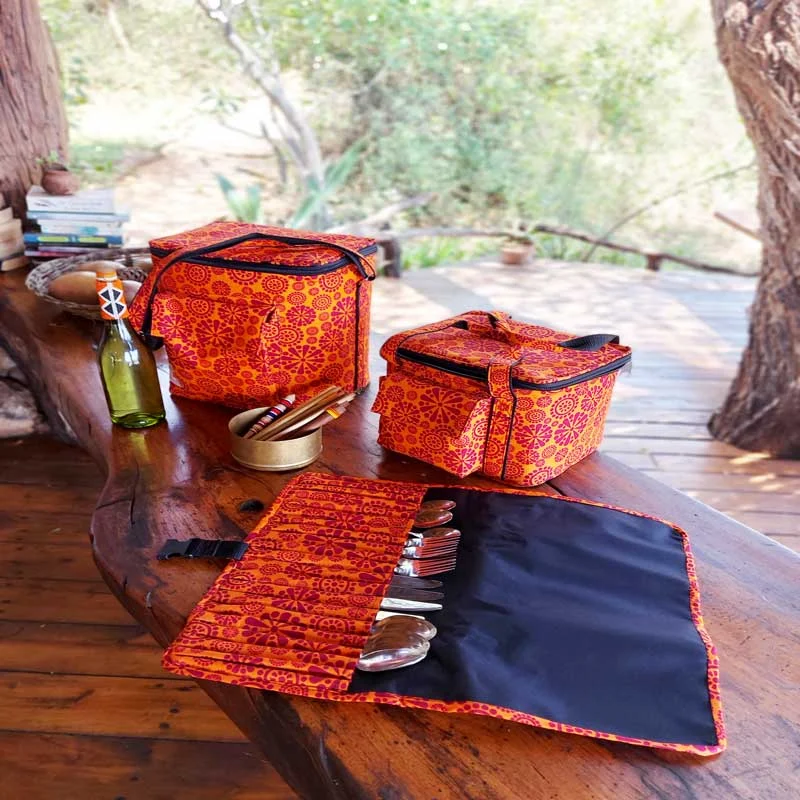 Two orange kanga cooler bags and a cutlery roll with steel cutlery on a wooden table on a deck