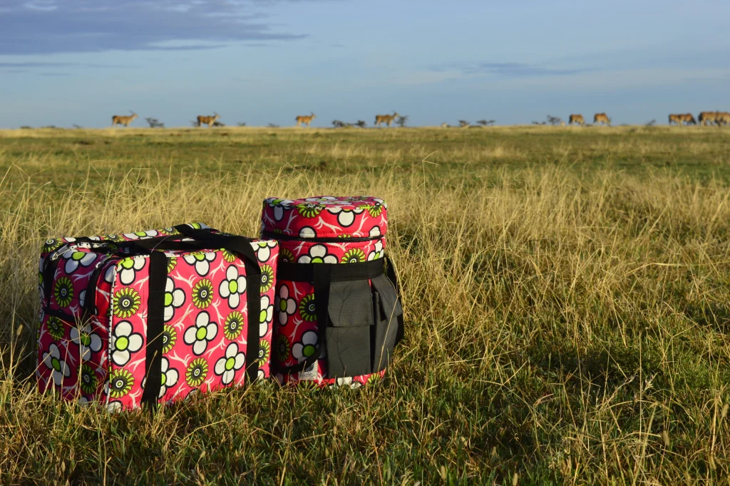 A pink flower-print kanga cooler bag and a round bottle cooler in long grass, with eland walking along the horizon