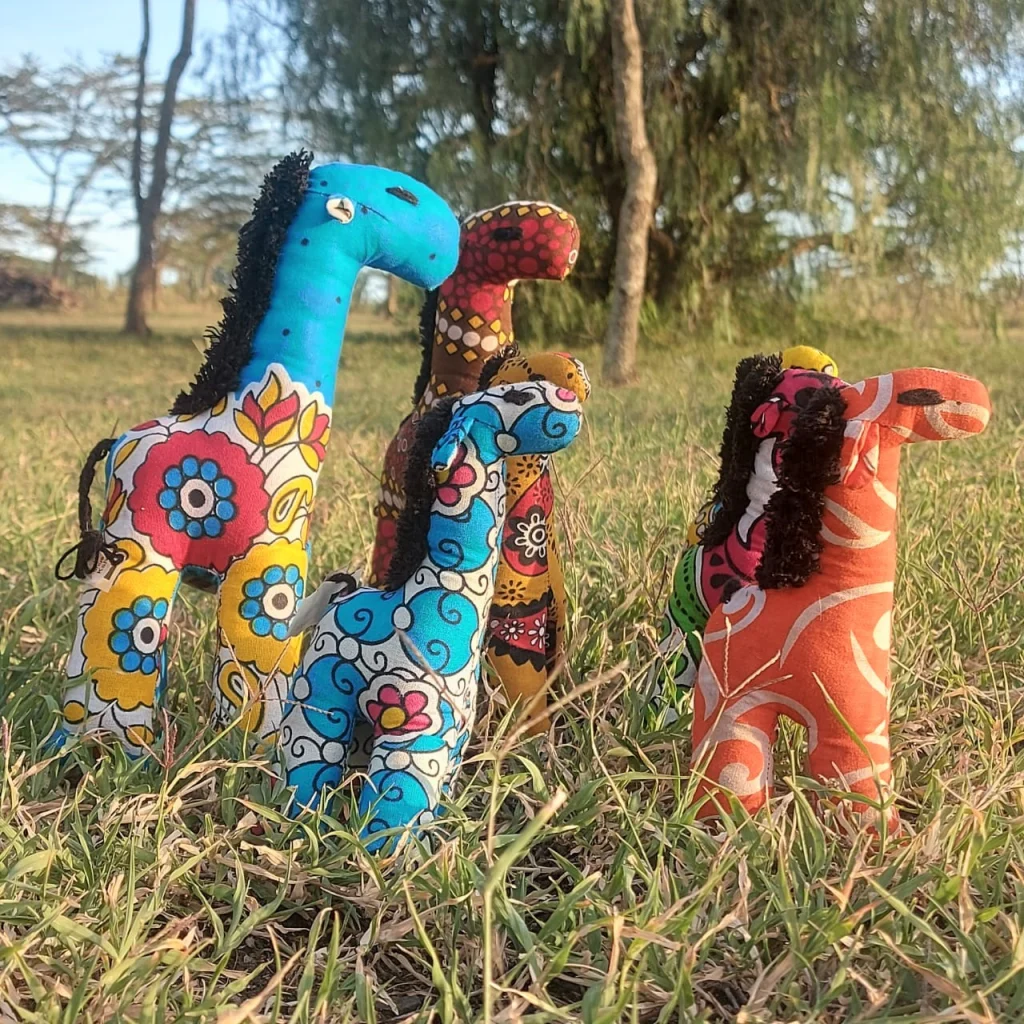 Five toy giraffes sewn from kanga prints standing in long grass under acacia trees