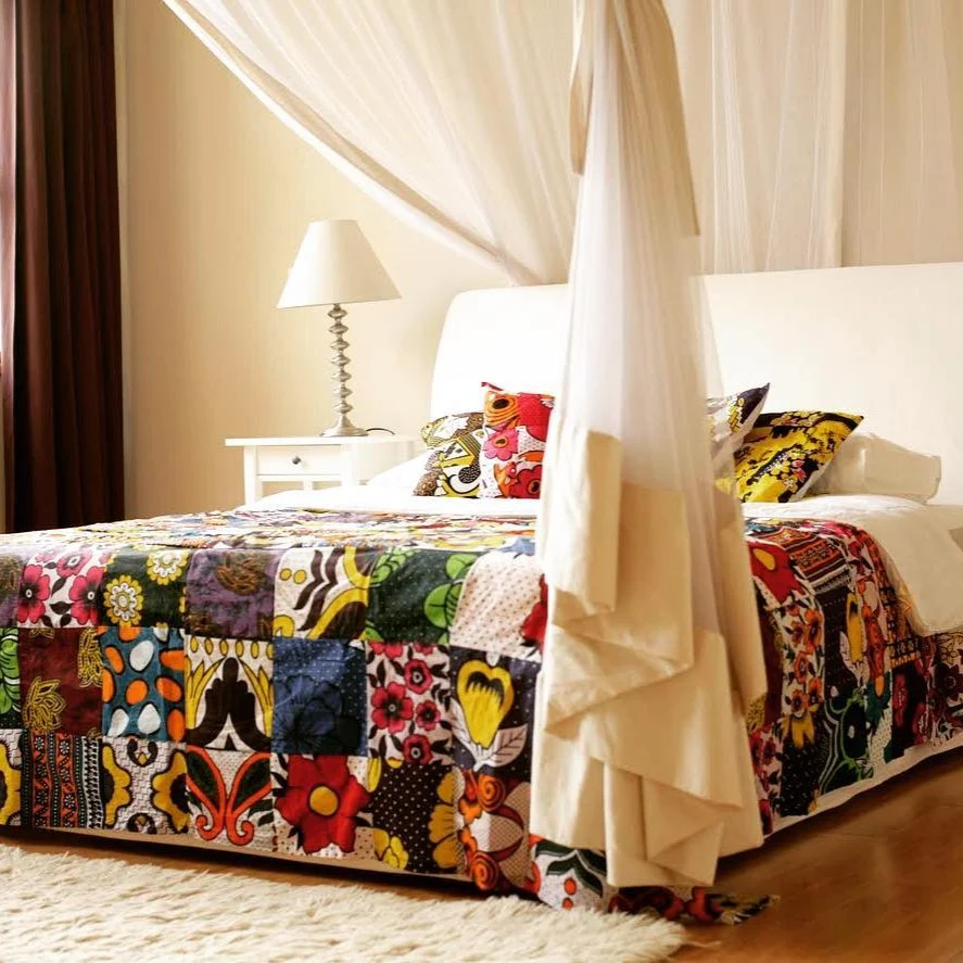 A bed with a colourful kanga patchwork bedspread and cushions under a white mosquito net
