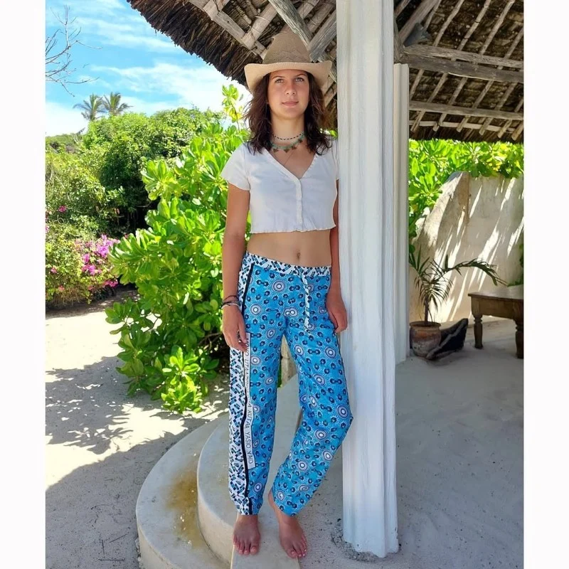 A young woman in blue kanga trousers and a white top leaning on a pillar under a thatched roof