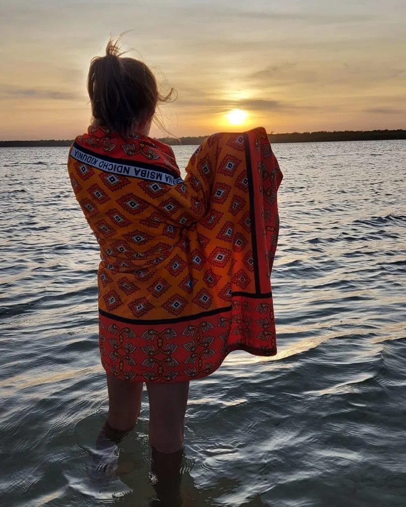 A woman standing in the sea at sunset wrapped in an orange and red kanga