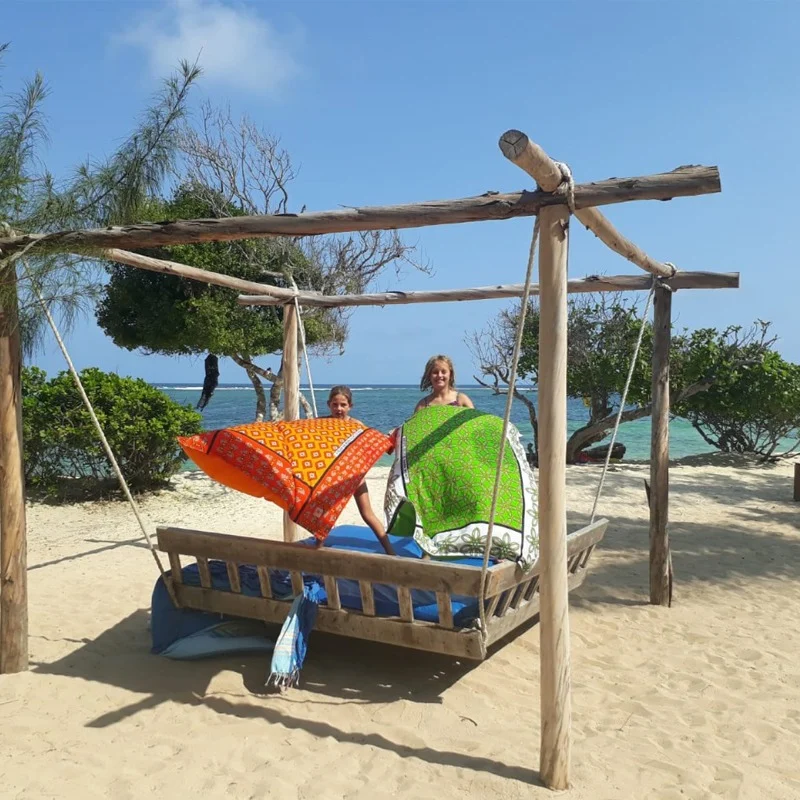Two kangas, one orange and one green, held up over a rope swing bed on the beach with the sea behind
