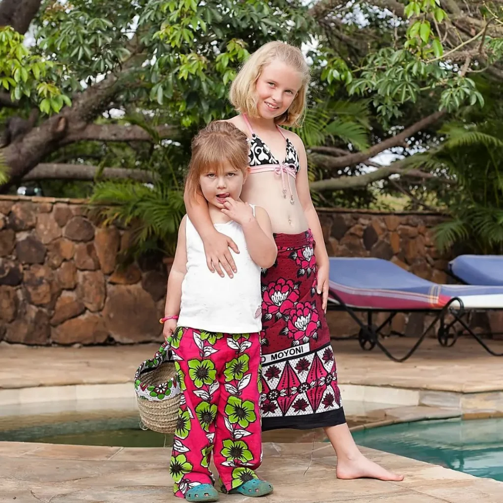 Two girls by a swimming pool, one in pink kanga trousers holding a small basket, the other in a red kanga wrap